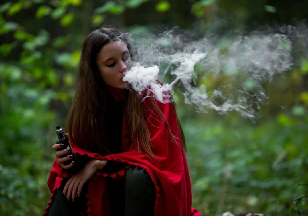 A person relaxing on a veranda, holding a disposable vape device with warm afternoon sun in the background, exhaling vapor.