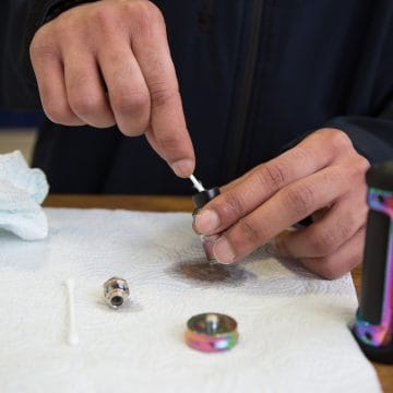A hand using a wooden toothpick to carefully dislodge lint and dry debris from the narrow airway of a disposable vape mouthpiece.
